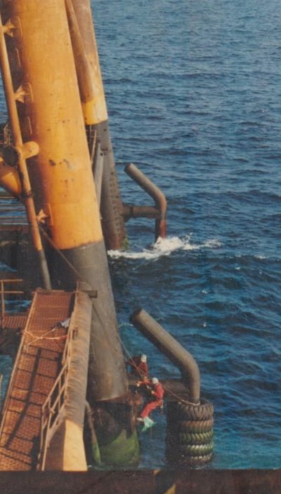 Two rope access technicians working down a platform leg above open water.