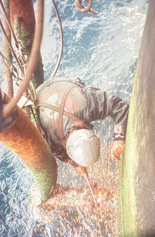 A rope access technician cutting steel above the sea, sparks falling.