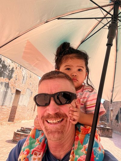 A man carrying a small child on his shoulders under a parasol.