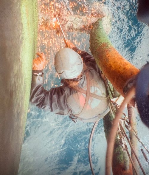 A rope access technician cutting steel high above open water, sparks falling into the sea.